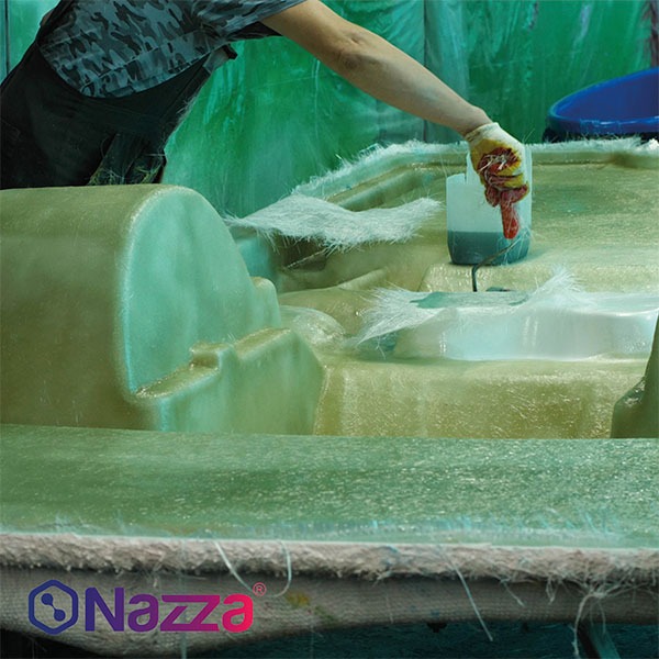 persona trabajando en un proceso de fabricación con resina de poliéster y fibra de vidrio. La persona está utilizando un guante de protección mientras vierte resina sobre una pieza que está siendo moldeada. La pieza está rodeada por fibras de vidrio que sobresalen de la superficie, lo que indica que está en una etapa de laminado o recubrimiento. El entorno está bien protegido con una pared verde, probablemente para evitar la dispersión de resina o material durante el proceso. El logo de Nazza aparece en la parte inferior izquierda, lo que sugiere que el proceso está relacionado con productos de esta marca.